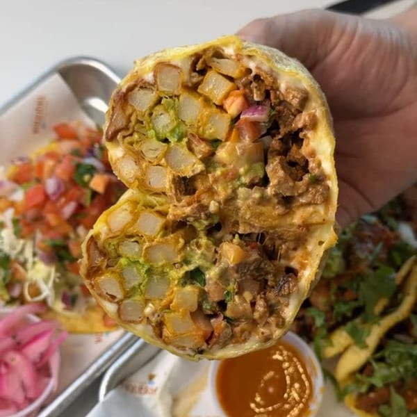Hand holding a Que Vida Taqueria burrito cut in half, revealing potatoes, meat, and guacamole.