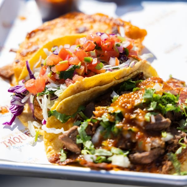 Close-up of three Que Vida Taqueria tacos with various fillings, salsa, and cilantro on a white tray.