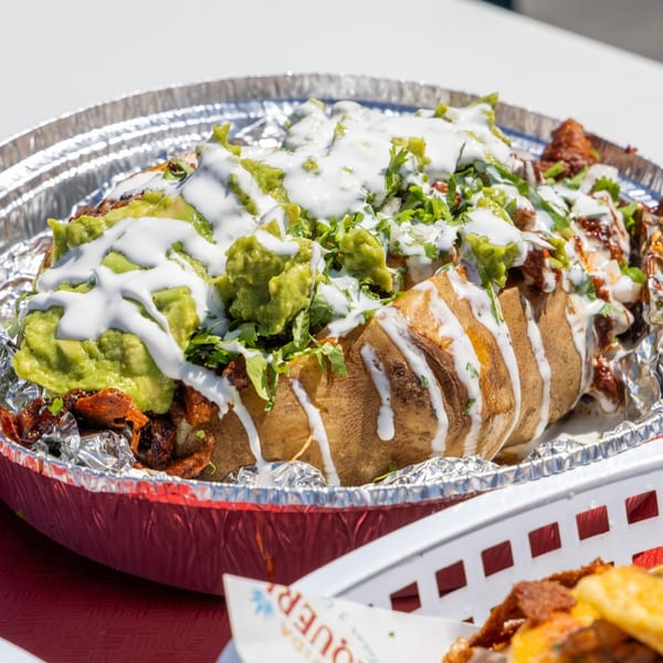 A large baked potato topped with guacamole, sour cream, and meat in a foil container.